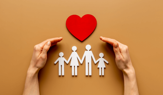The image shows a cut-out paper family of four (two adults and two children) holding hands, positioned below a red heart symbol. Two hands are placed around the family, appearing to protect or embrace them. The background is a solid tan color. The image conveys themes of family care, love, and protection, and explore top benefits of having a life insurance policy.
